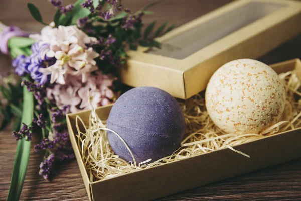 放松洗浴用品作为礼物的概念 两个色彩艳丽的浴弹放在一个褐色纸盒里 背景是木制桌子上装饰着粉红色和紫色花朵 — 图库照片