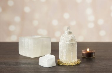 Doğal olarak oyulmuş mineral taşından yapılmış Selenit kulesi temizlik özelliklerinde iyileşme gösterir. Mumluk, çiğ ve oyulmuş Selenit kule koyu ahşap masa, turuncu bokeh arkaplan.