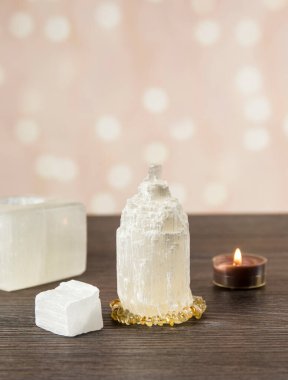 Doğal olarak oyulmuş mineral taşından yapılmış Selenit kulesi temizlik özelliklerinde iyileşme gösterir. Mumluk, çiğ ve oyulmuş Selenit kule koyu ahşap masa, turuncu bokeh arkaplan.