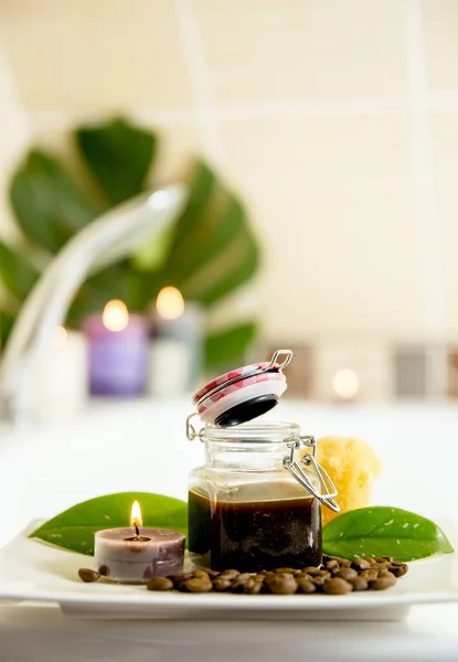 Coffee scrub mixture in glass jar for using many purposes: face, hair, body mask and scrub concept, indoors in home bathroom, candles lit flames on the background.