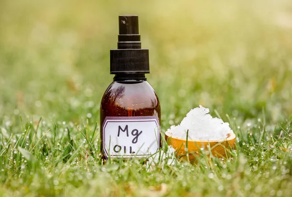 Magnesium Chloride Flakes on brown wooden cup with Magnesium vitamin oil in spray bottle in grass outdoors with blurred bokeh green background.