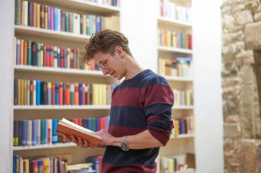 Gözlüklü ve bukleli genç bir öğrenci kütüphanede kitaplığın yanında kitap okuyor..