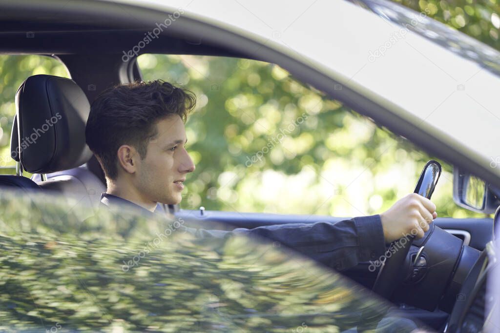 Profile of a young handsome driver sitting in his car with hands at the wheel, the side window half open and with light reflections                     