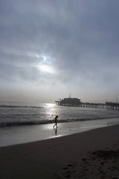 Shadowy picture of a little girl running through the sand  at Santa Monica Beach, in the background sunset and pier, sky is cloudy but bright