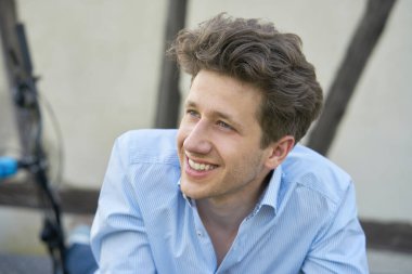 Kahverengi bukleli ve üç günlük sakallı genç bir adamın portresi. Mavi bir gömlek içinde oturuyor, beyaz dişleriyle gülümsüyor.