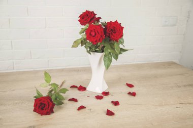 Masada beyaz vazo ve gül yaprakları olan kırmızı güller.