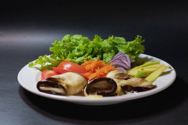 Sağlıklı vejetaryen yiyecekler, beyaz tabakta ve siyah arka planda, tabağın patateslerden, havuçlardan, domateslerden, marullardan ve avokadodan oluştuğunu görebiliyoruz.