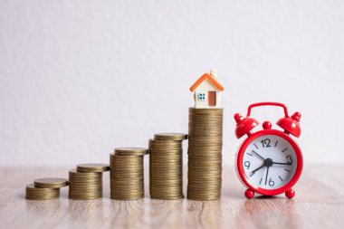 Üzerinde ev olan bozuk para yığınının yanına yerleştirilmiş kırmızı alarm saati. Zamanı iyi kullanma ve bu süre zarfında konut işine yatırım yapma fikri risk altında olabilir..