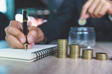 Bir iş adamının eli cam bir şişeye bozuk para koyar. Hand bir gelir ve gider hesabı yazmak üzereydi. Finansal muhasebe kavramları. İş başarısı için varlıkları hesapla.