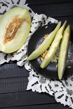 Olgun kavun dilimlerinin üst görüntüsü siyah tabakta ve kavunun yarısı beyaz el yapımı peçetenin üzerinde ahşap masada yatıyor.