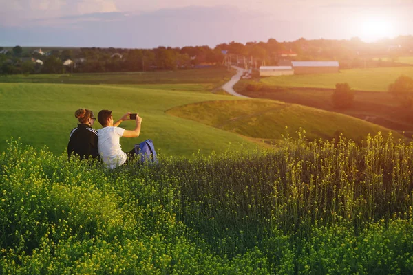 Young loving couple sits on a hill with phone on the green field background — Stock Photo, Image