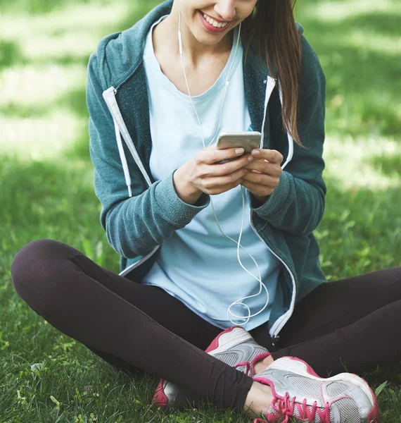Portrait of young and sporty woman in sportswear sitting with the smartphone on the grass in park — Stock Photo, Image
