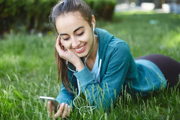 Portrait of young and sporty woman in sportswear Lies with the smartphone on the grass in park — Stock Photo, Image