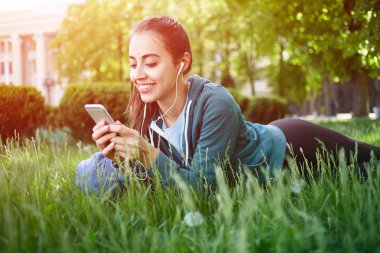 Spor giyim genç ve sportif kadın portresi ile smartphone Park çimenlerin üzerinde yatıyor