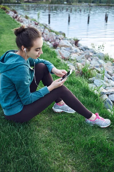 Portrait of young and sporty woman in sportswear sits with the smartphone on the grass in park — Stock Photo, Image