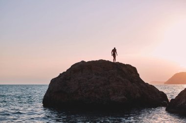 gün batımında denizin içinde büyük bir kayanın üzerinde bir adam duruyor
