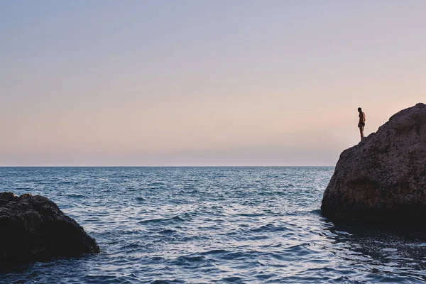 gün batımında denizin içinde büyük bir kayanın üzerinde bir adam duruyor