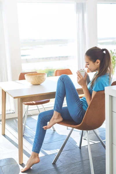 En ung kvinne sitter på kjøkkenet om morgenen med en kopp kaffe – stockfoto
