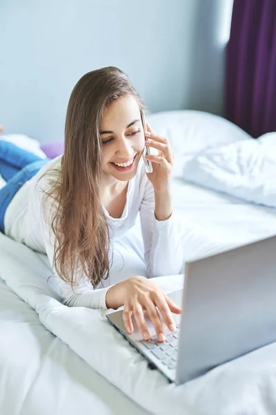 En ung smilende kvinne med bærbar PC som bruker telefon – stockfoto