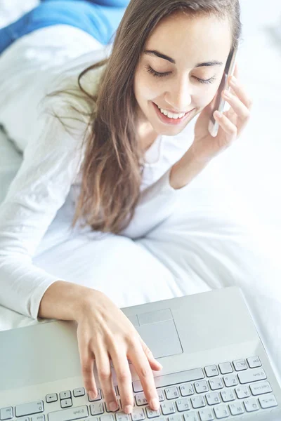 En ung smilende kvinne med bærbar PC som bruker telefon – stockfoto