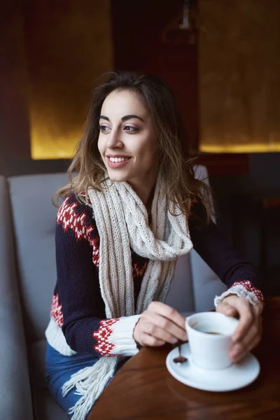 Mujer sonriente vestida con un suéter y bufanda en la cafetería con taza de café — Foto de Stock