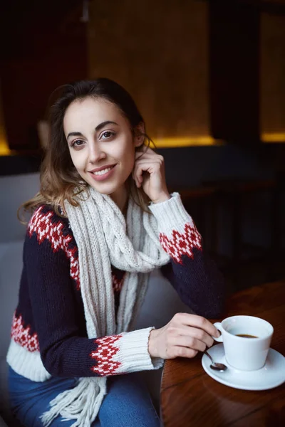 Mujer sonriente vestida con un suéter y bufanda en la cafetería con taza de café — Foto de Stock