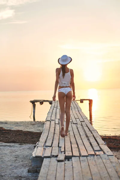 Kaunis seksuaalinen nainen valkoisessa hatussa ja bikinit puulaiturilla merta ja auringonlaskua vastaan — kuvapankkivalokuva