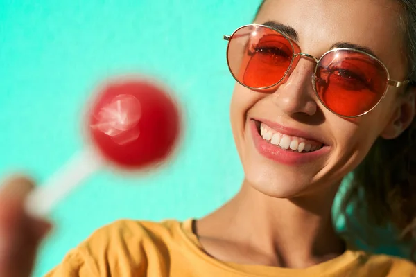 Gros plan portrait d'une jeune femme élégante souriant à la caméra, montrant une grosse sucette rouge sur fond bleu . — Photo