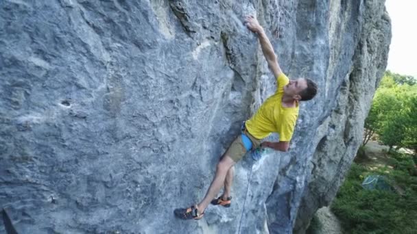 Vide angle view of man rock climber in yellow t-shirt, climbing on a cliff, on tough sport route, resting and chalking hands. outdoors rock climbing and active lifestyle concept — Stock Video