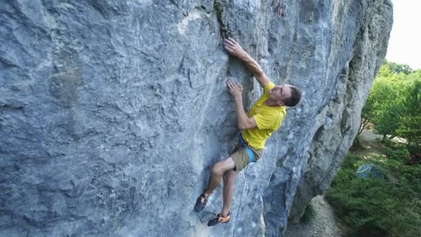 Vide angle view of man rock climber in yellow t-shirt, climbing on a cliff, searching, reaching and gripping hold. outdoors rock climbing and active lifestyle concept — Stock Video