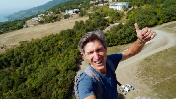 Fotografo uomo sorridente cattura selfie video in piedi sul bordo della scogliera su montagne, cielo e sfondo marino . — Video Stock