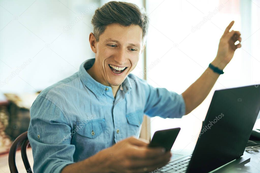 Portrait of an excited happy handsome man using phone and laptop computer at home