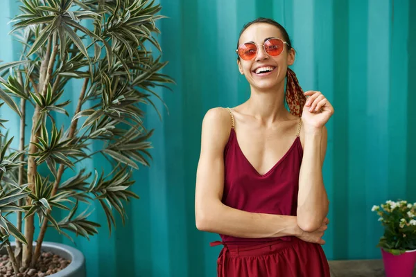 Giovane bella donna in occhiali da sole alla moda e vestito rosso elegante ridendo alla macchina fotografica su sfondo blu con grandi piante in vasi ai lati. — Foto Stock