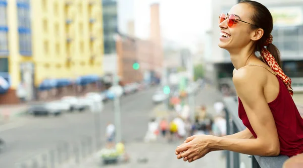 Giovane donna sorridente in occhiali da sole alla moda e vestito rosso elegante godendo della vista sulla città dal balcone. — Foto Stock