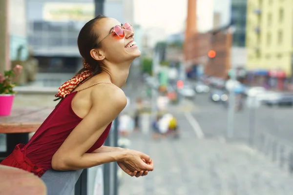 Joven mujer sonriente con gafas de sol de moda y ropa roja con estilo que disfruta de la vista de la ciudad desde el balcón.. —  Fotos de Stock