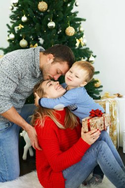 Genç bir aile Noel 'i evde kutluyor, güzel bir Noel ağacının yanında oturuyor, gülüyor, sarılıyor ve hediyeler veriyor.