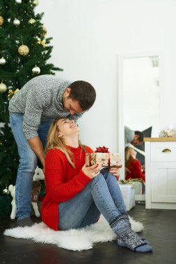 Genç bir adam Noel hediyesiyle sevgilisine sürpriz yapıyor. Arka planda Noel ağacı var..