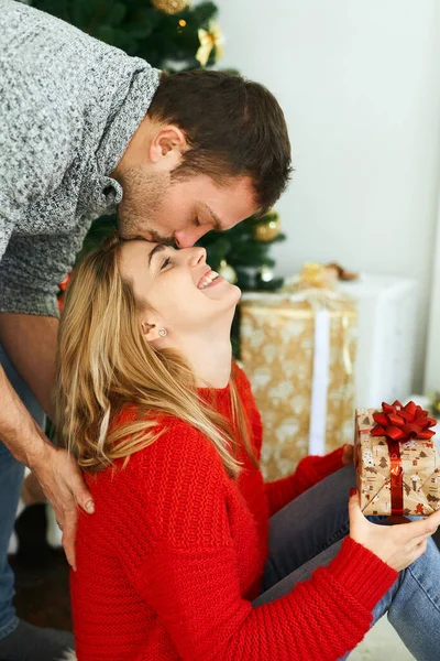 Young man presenting Christmas gift his girlfriend at home with christmas tree on background.