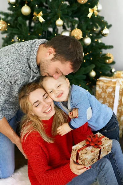 Ung familie feirer jul hjemme, sitter nær vakkert juletre, ler, omfavner og gir gaver – stockfoto
