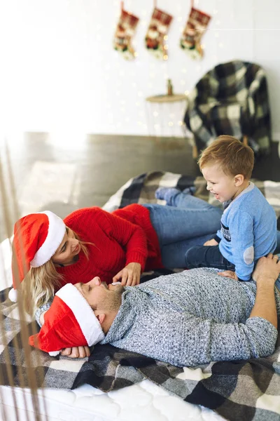 Happy young couple in Christmas Santa hats lying on bed with their son with Christmas decoration on background. – stockfoto