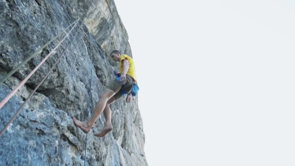 Sonriente hombre satisfecho escalador de roca con los pies desnudos, vistiendo en camiseta amarilla, bajando después de subir por un acantilado. al aire libre escalada en roca y concepto de estilo de vida activo — Vídeo de stock