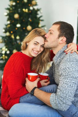 Birbirine aşık romantik bir çift, arka planda yılbaşı ağacı olan, kahve bardaklarını kucaklıyor, öpüyor ve tutuyorlar..