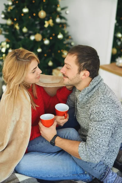 Romanttinen pari rakastunut peitetty lämpimällä pehmeällä peitolla, syleilemällä ja pitämällä kuppeja kahvia kotona joulukuusi taustalla . — kuvapankkivalokuva