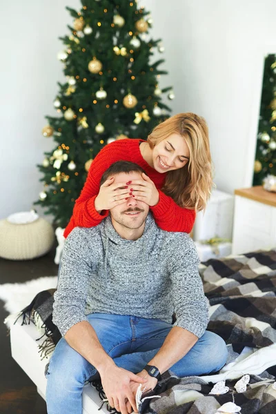Yong nainen sulkemalla hänen poikaystävänsä silmät kädet, istuu sängyllä lähellä joulukuusi kotona . — kuvapankkivalokuva