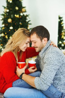 Birbirine aşık romantik bir çift, arka planda Noel ağacı olan kahve bardaklarını kucaklıyor ve ellerinde tutuyorlar..