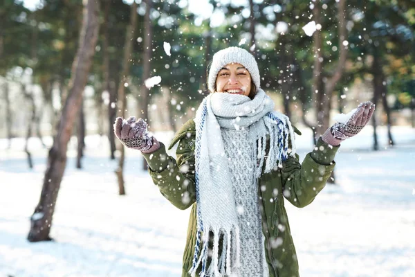 Zimowy portret zabawnej kobiety w wełnianym kapeluszu i długim ciepłym szaliku rzucającej śniegiem w zimowym parku, latające płatki śniegu. — Zdjęcie stockowe
