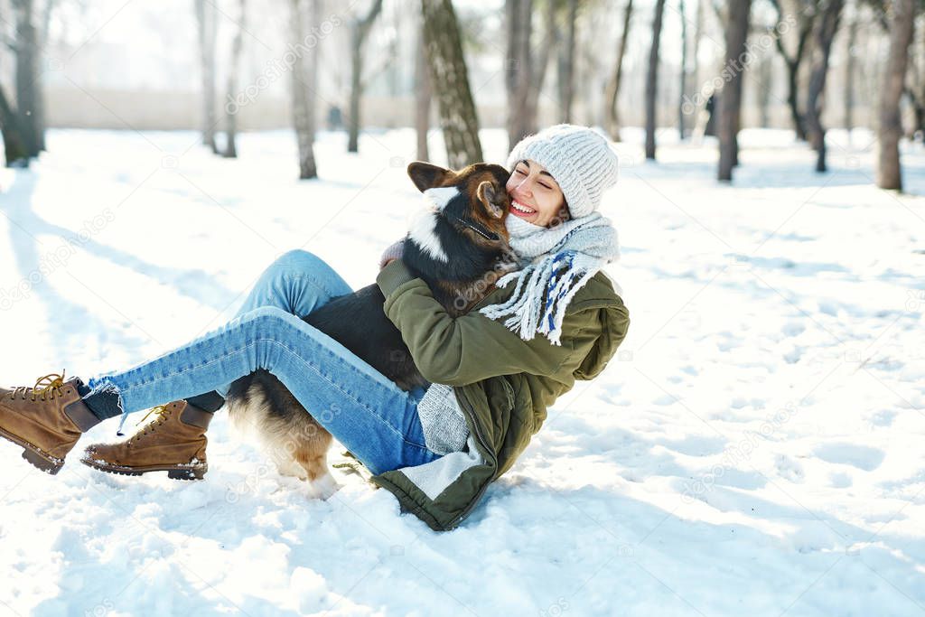 Young happy woman in woolen hat and long warm scarf hugs her pet in snowy winter park at frozzy sunny day