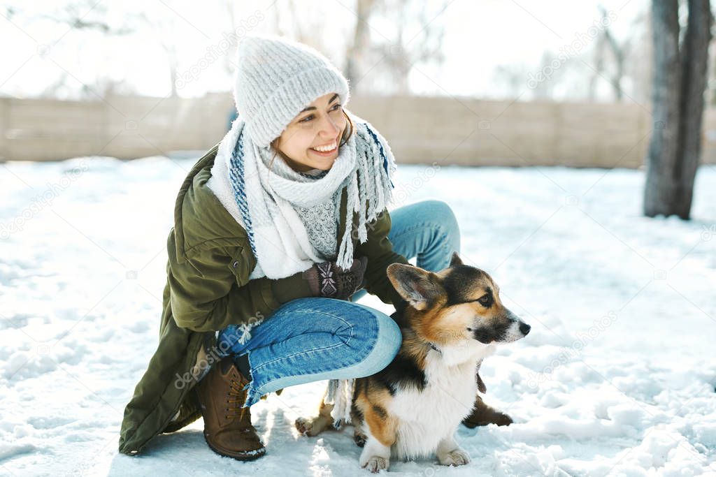 Young happy woman in woolen hat and long warm scarf stroking her pet and having fun in snowy winter park at frosty sunny day