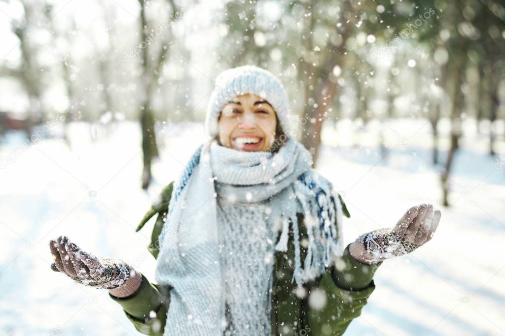Winter portrait of funny woman in woolen hat and long warm scarf throwing snow in winter park, flying snowflakes.
