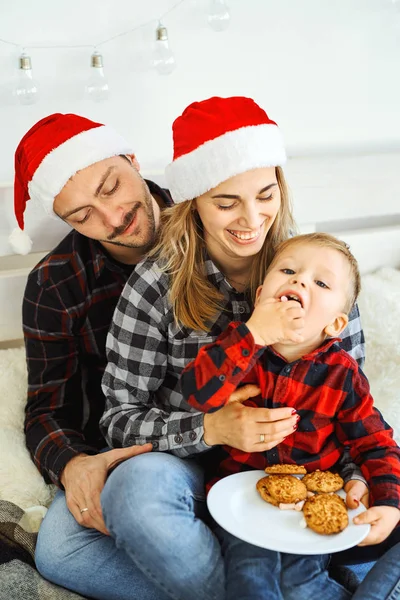 Julefamilie i julenissehatter. Pappa, mor og sønn tilbringer tid sammen, spiser småkaker og klemmer i sengen hjemme . – stockfoto